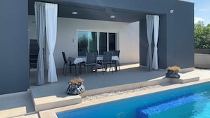 Een modern terras buiten met een zwembad, inclusief een eethoek met stoelen en een tafel, en twee decoratieve plantenbakken.