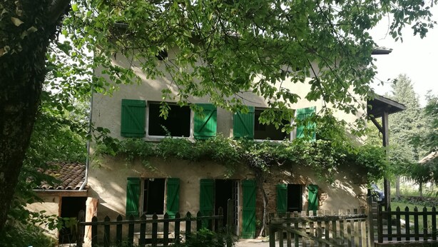 Un edificio de dos pisos con contraventanas verdes, una valla de madera y un árbol en primer plano.