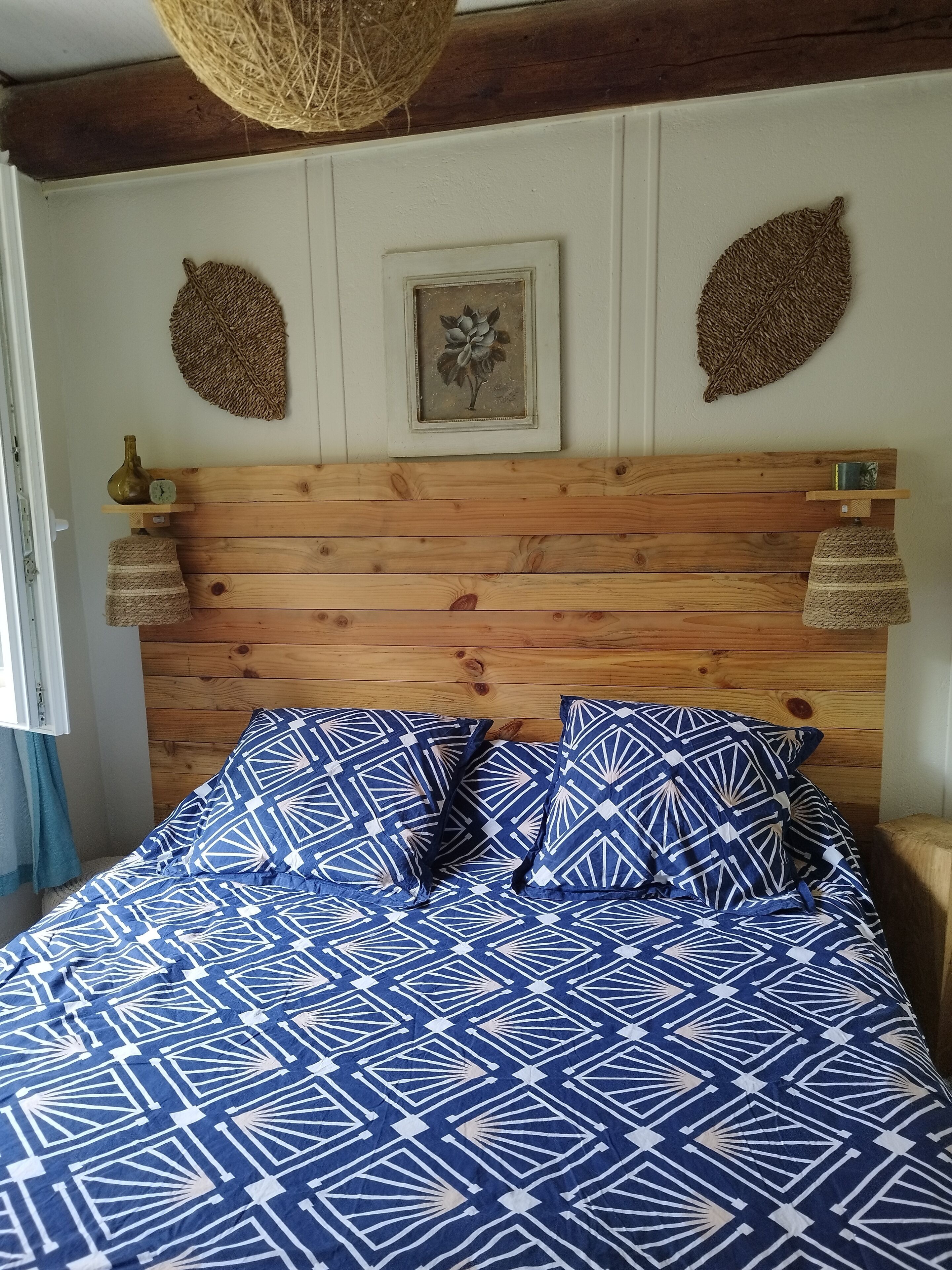 Une chambre à coucher avec une tête de lit en bois, un lit recouvert d’une literie à motifs bleus, un tableau encadré et deux lampes fixées au mur.