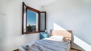 Ein Schlafzimmer mit großem Fenster, einem Bett mit aufgerollten Handtüchern, einem Nachttisch und einem hölzernen Kopfteil.