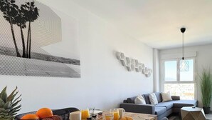 A modern living room with a gray sofa, a coffee table with fruits and drinks, a TV, and a large window with a view. - ONE7 Suite – The sunrise penthouse (Estepona)