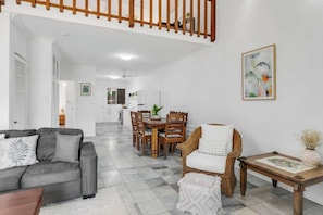 A living room with a sofa, a dining table with chairs, a wicker chair, a small table with a vase, and a staircase.