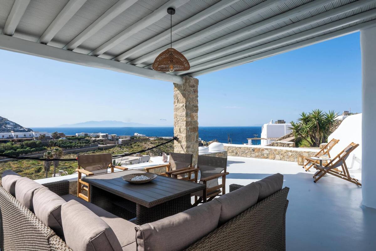 A terrace with wicker furniture, a stone pillar, and a view of the sea and buildings.