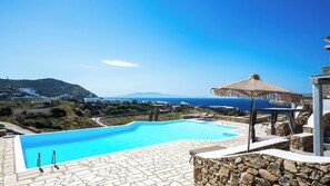 A clear blue swimming pool with stone tiles, a thatched umbrella, and a view of the sea and distant hills.