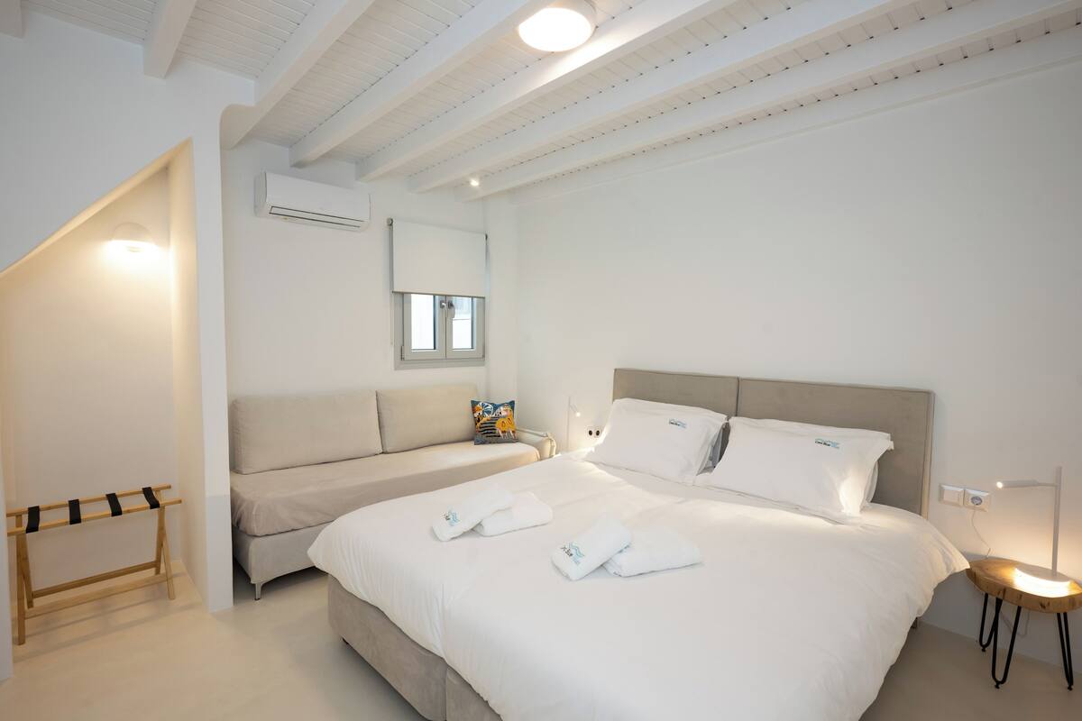 A modern hotel room with a large bed, a smaller sofa, a wooden rack, and a bedside table with a lamp.