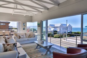 Una sala de estar moderna con un ventanal grande, un sofá, una mesa de centro y vistas a una casa costera.