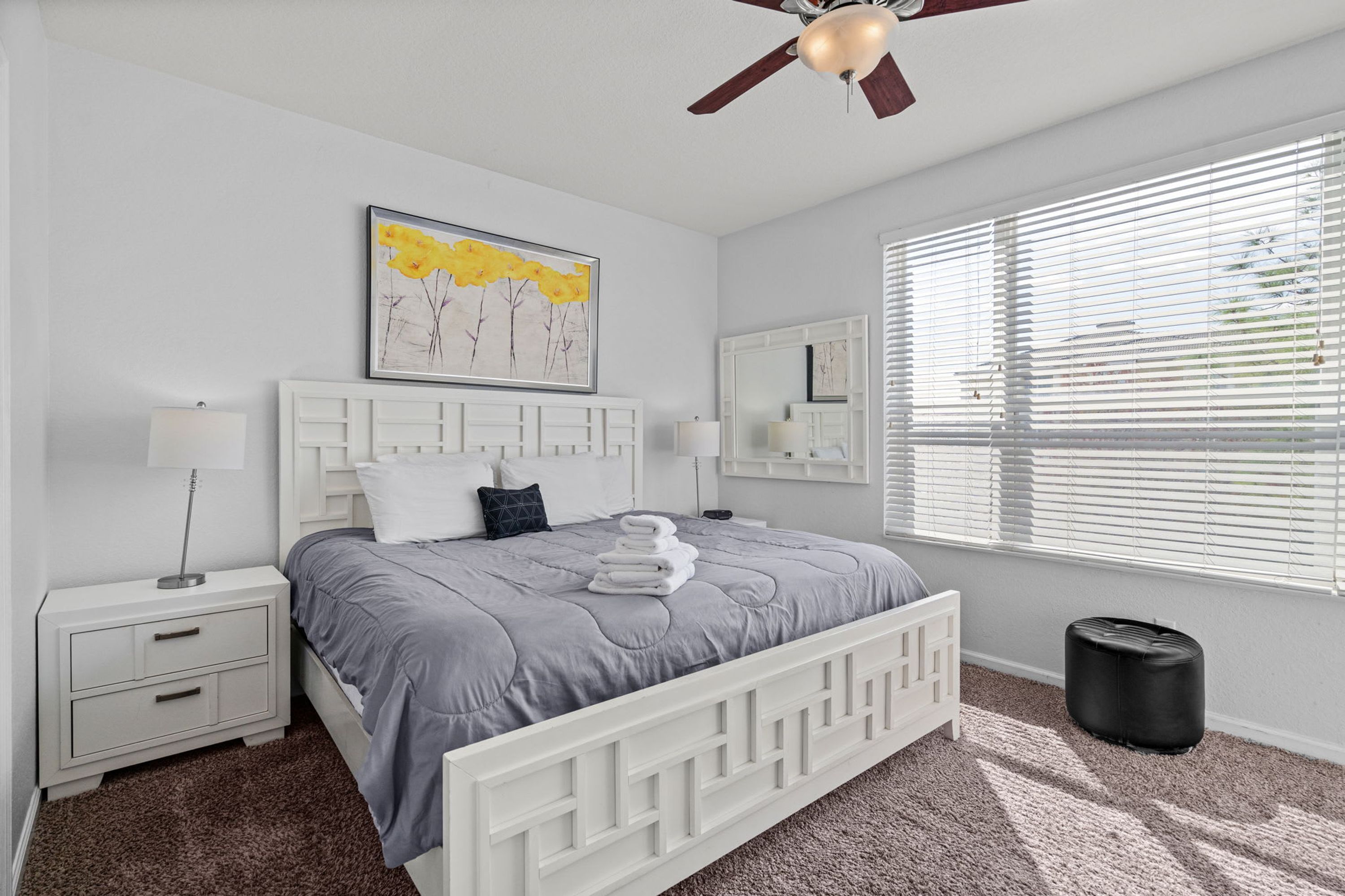 A bedroom with a white bed, a ceiling fan, a window with blinds, a bedside table with a lamp, and a framed picture on the wall.