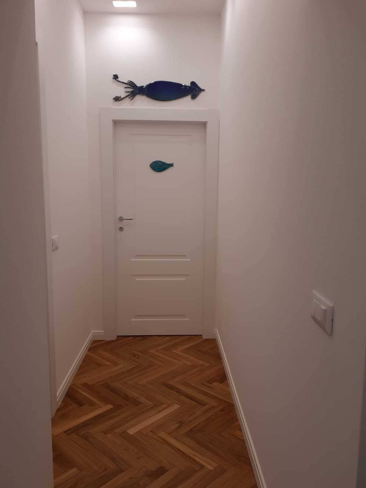 Un corridoio stretto con pavimento in parquet a spina di pesce, una porta bianca decorata con un motivo a forma di pesce e una lampada a soffitto.
