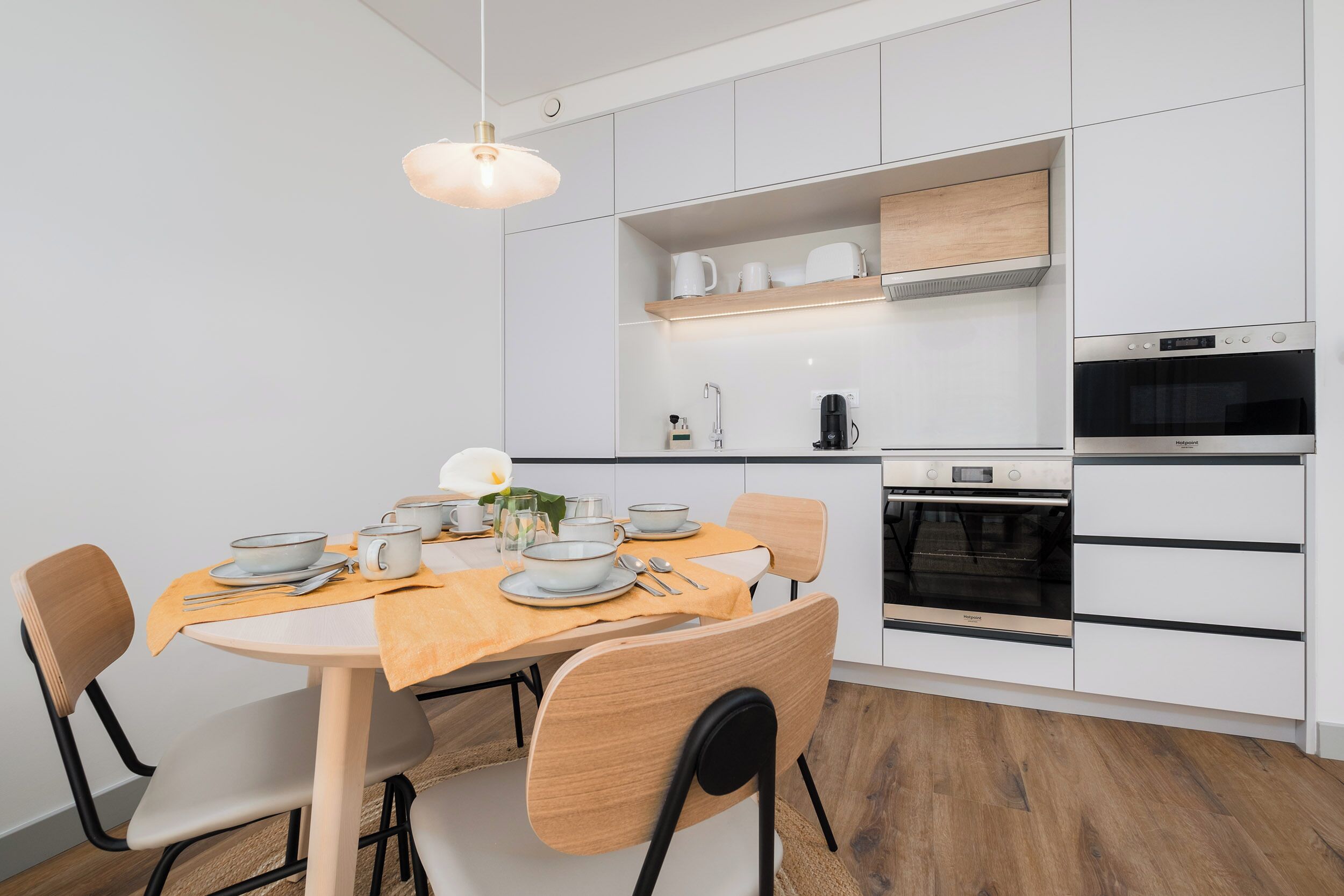 Una cocina moderna con armarios blancos, horno empotrado y comedor con mesa y sillas de madera.