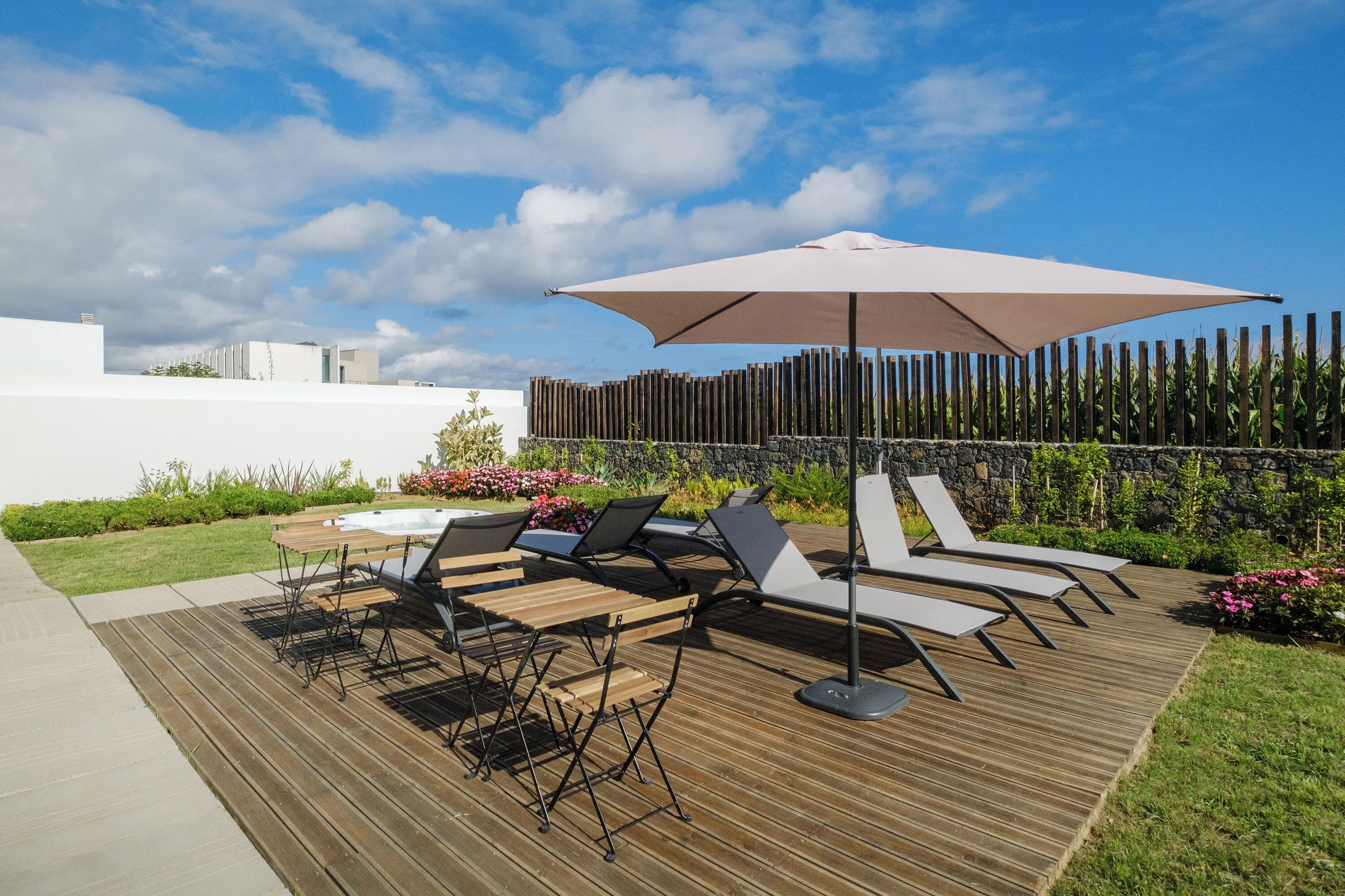 A deck with lounge chairs and a table, a large umbrella, and a stone wall with plants.