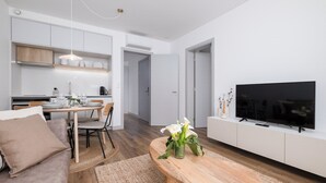 A modern living room with a wooden dining table, a sofa, a TV, and a kitchen area with cabinets and a hanging light.