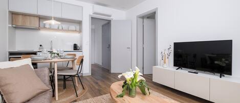 Ett modernt vardagsrum med ett matbord i trä, en soffa, en tv och en köksdel med skåp och en hängande lampa.