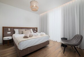 Una habitación de hotel moderna con una cama grande, una mesita redonda, una silla y un ventanal con cortinas.