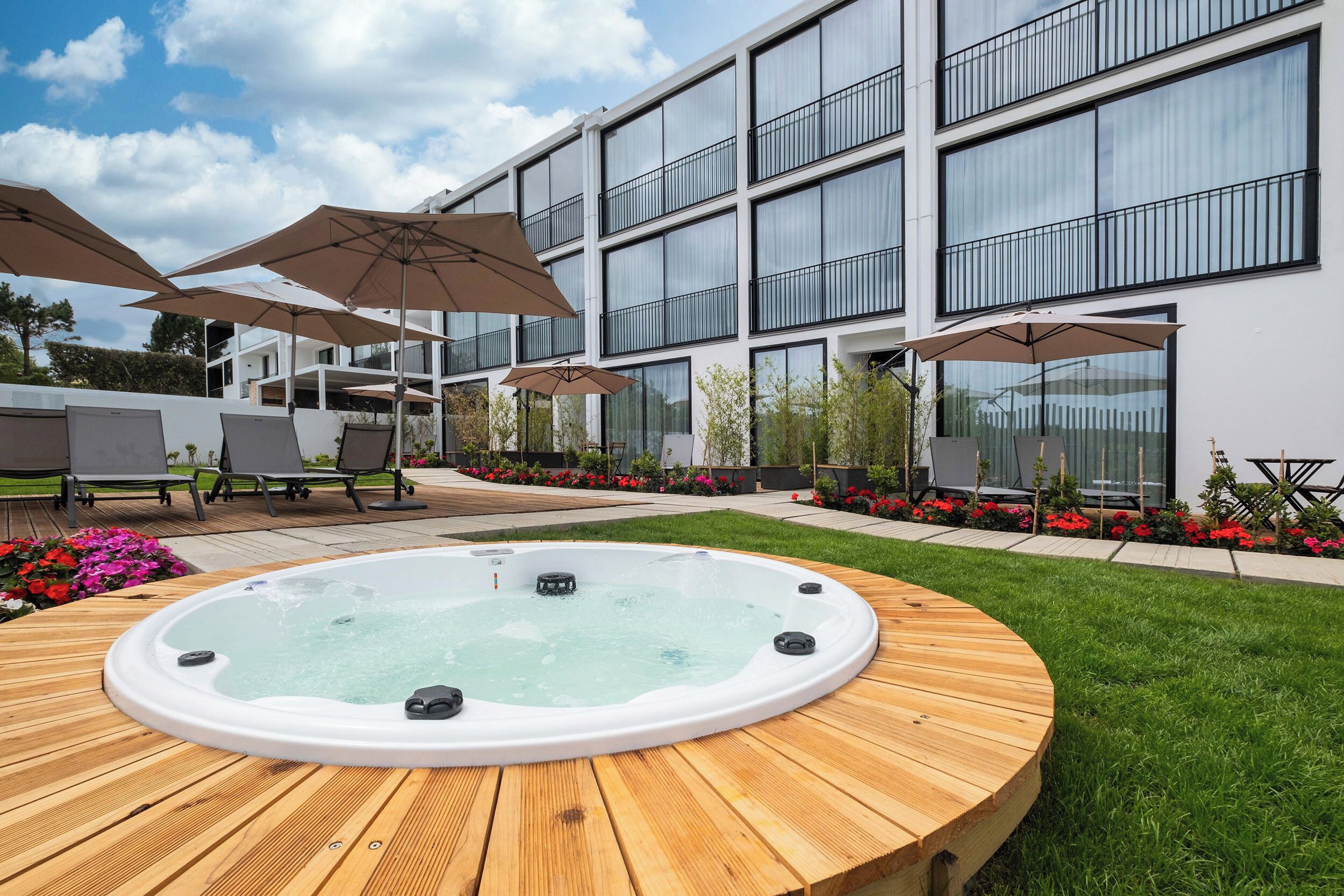 Un edificio moderno con ventanas amplias, un jacuzzi en una terraza de madera, mobiliario exterior con sombrillas y un jardín bien cuidado.