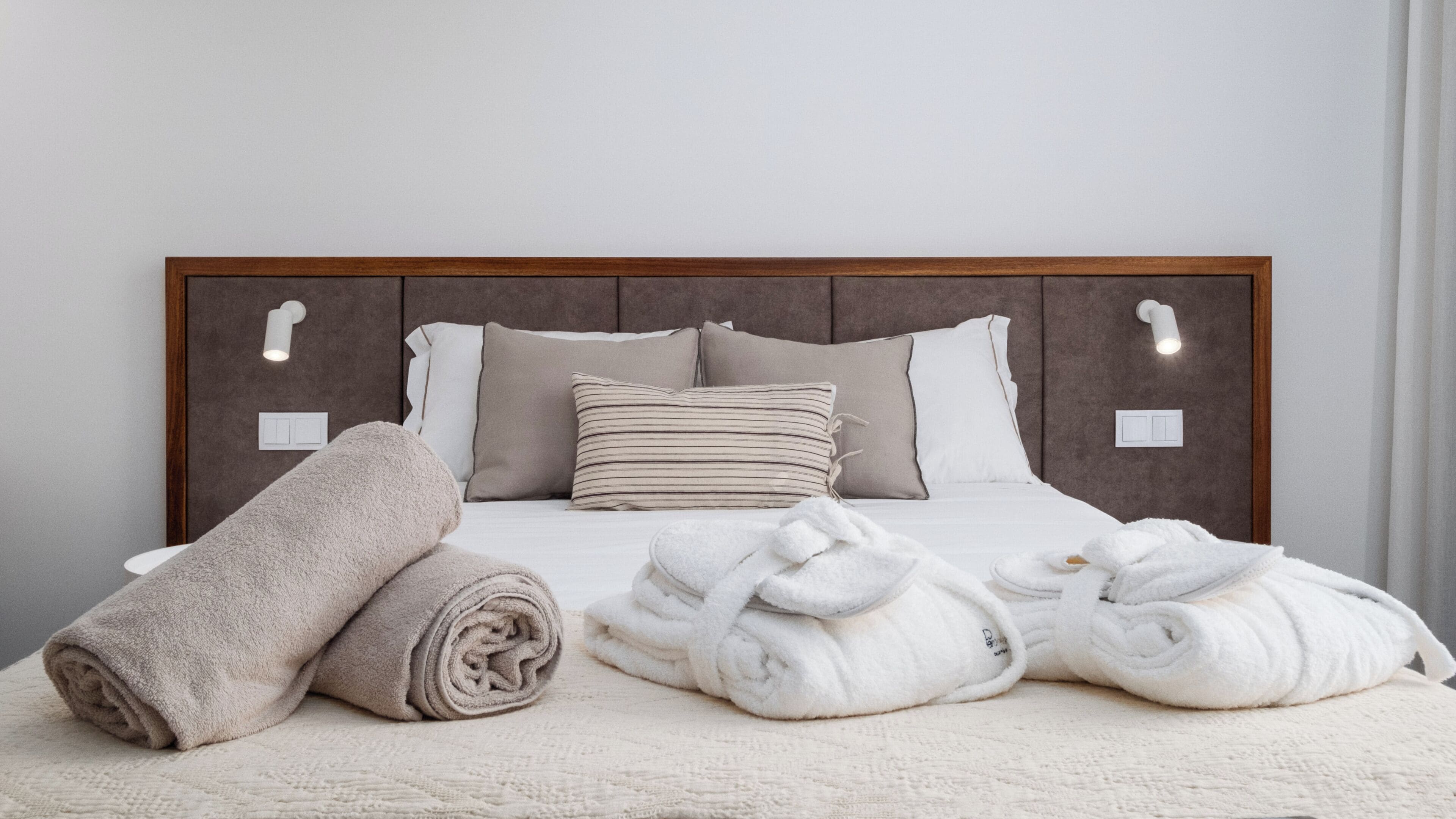 Ein ordentlich bezogenes Bett mit einem Holzkopfteil, zwei Nachttischlampen sowie einem zusammengerollten Handtuch und zwei Bademänteln auf dem Bett.