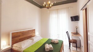 A hotel room with a wooden headboard bed, a green bedspread, two rolled towels, a desk with a chair, a flat-screen TV, and a window with sheer curtains. - Visitsicilia Donn'Anna 3 (Castellammare del Golfo)