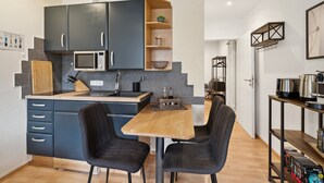 A compact kitchen with dark cabinets, a microwave, and a sink. There are two chairs at a wooden table, and a bookshelf with books and a kettle. - Charysma: Aurum Suite | modern & elegant design | parking lot | terrace (Hameln)