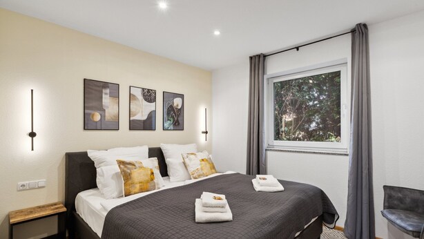 A modern bedroom with a large bed, a nightstand, a chair, and a window with curtains. - Charysma: Aurum Suite | modern & elegant design | parking lot | terrace (Hameln)