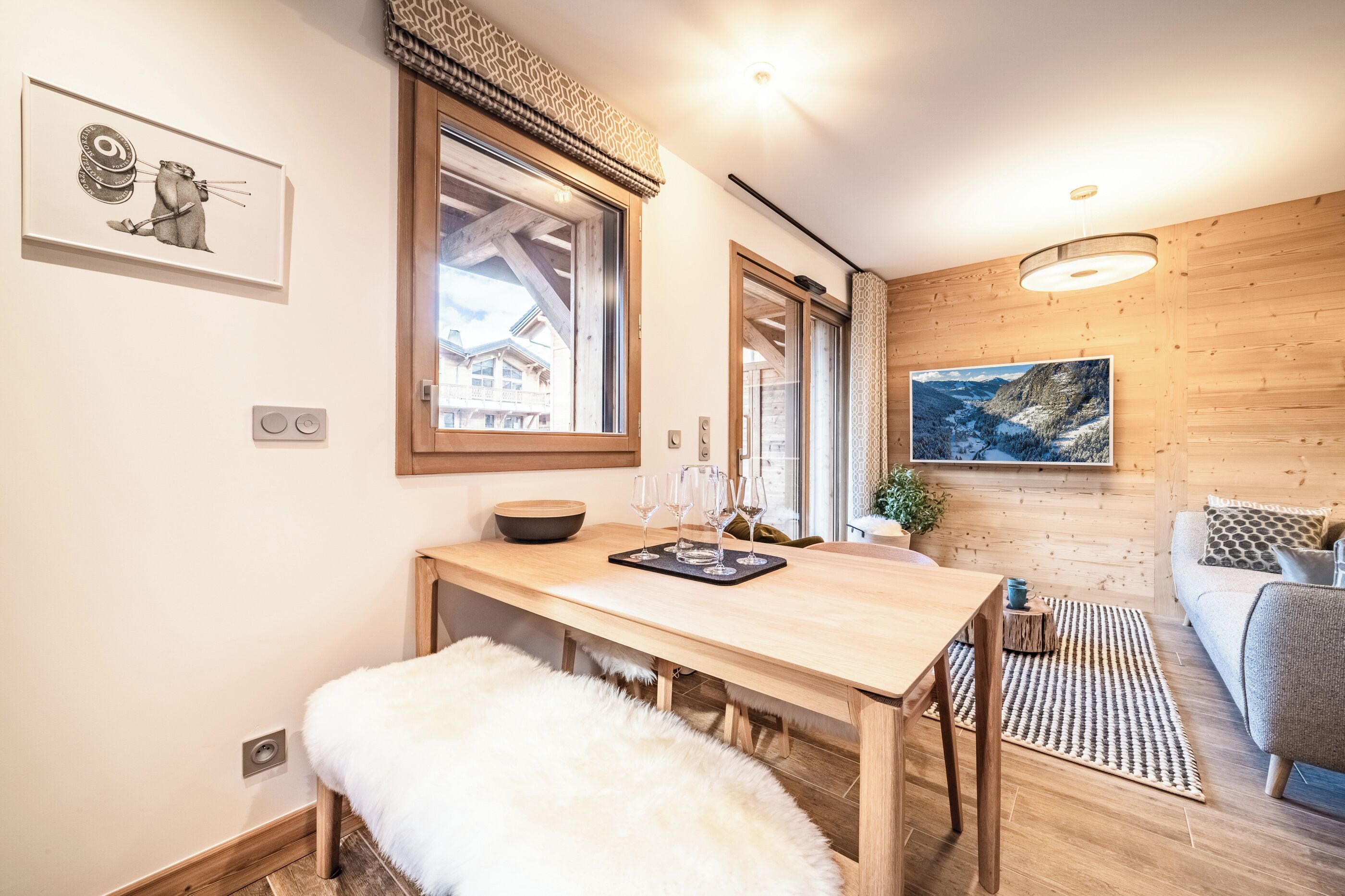 Une pièce chaleureuse avec une table à manger en bois, un banc blanc et moelleux, une grande fenêtre et un tableau accroché au mur.