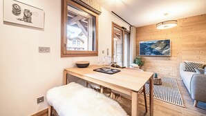 Een gezellige kamer met een houten eettafel, een zachte, witte bank, een groot raam en een schilderij aan de muur.
