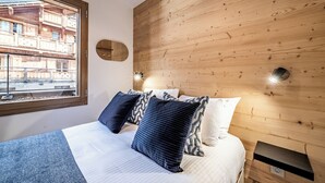 A bedroom with a wooden headboard, a bed with white and blue bedding, and a window with a view of a building. - Fyndor - Apartment located in center of Morzine (Morzine)