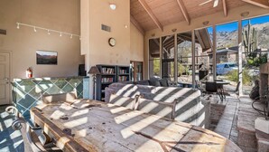 Ein geräumiger Innenbereich mit Holzboden, einem großen Tisch und einer Couch. Es gibt Bücherregale und ein Fenster mit Blick ins Freie.