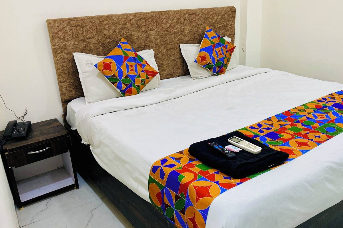 A neatly made bed with a white duvet, two colorful geometric pillows, a black blanket with a remote, and a phone on a bedside table.