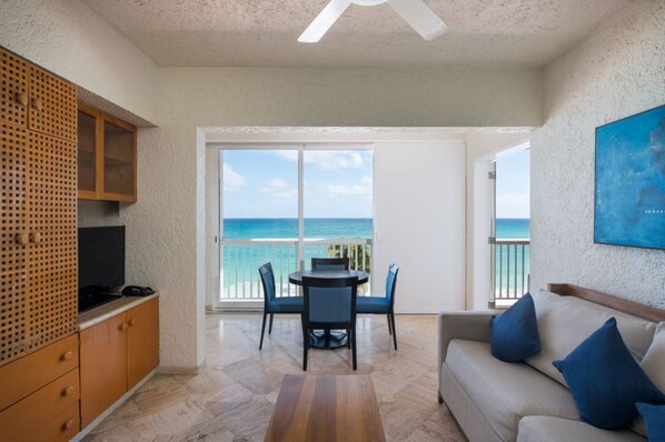 A room with a balcony overlooking the sea, a dining table with chairs, a sofa, and a TV on a wooden cabinet.