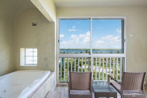 Une chambre dotée d’une grande baignoire, d’un balcon avec des fauteuils en osier et offrant une vue sur un plan d’eau et la verdure.