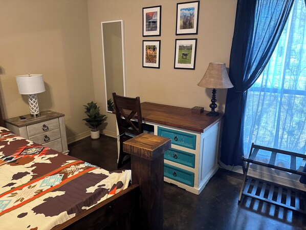 A bedroom with a bed, a desk, a chair, a dresser, a mirror, a lamp, a potted plant, and framed pictures on the wall.