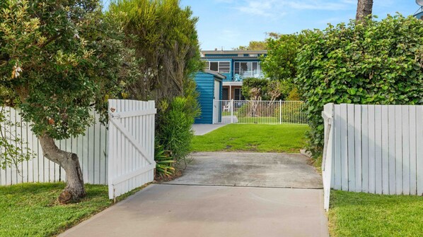 A residential property with a paved driveway leading to a house with a blue exterior. There is a white picket fence surrounding the property.