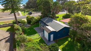 A single-story house with a blue exterior, surrounded by lush greenery and a clear view of the ocean. - Surfrider Studio - Pet Friendly - 50m to Beach - Book 3 Nights, Pay for 2 until 24 September 2026 (Culburra Beach)