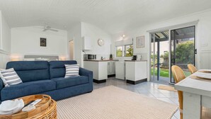 A modern living room with a blue sofa, a kitchen with white cabinets, and a sliding glass door leading to a garden. - Surfrider Studio - Pet Friendly - 50m to Beach - Book 3 Nights, Pay for 2 until 24 September 2026 (Culburra Beach)