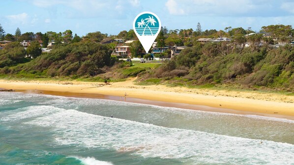 Vista costera con playa, olas del océano y zona residencial con casas y árboles al fondo.