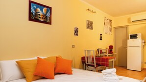 Una camera d'albergo con un letto, cuscini, una sedia, un tavolo con tovaglia rossa, un frigorifero e una parete con quadri.