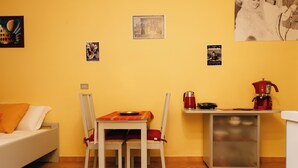 Una camera compatta con un letto, un tavolo da pranzo con sedie, un angolo cottura con una macchina del caffè e uno scaffale con vari oggetti.