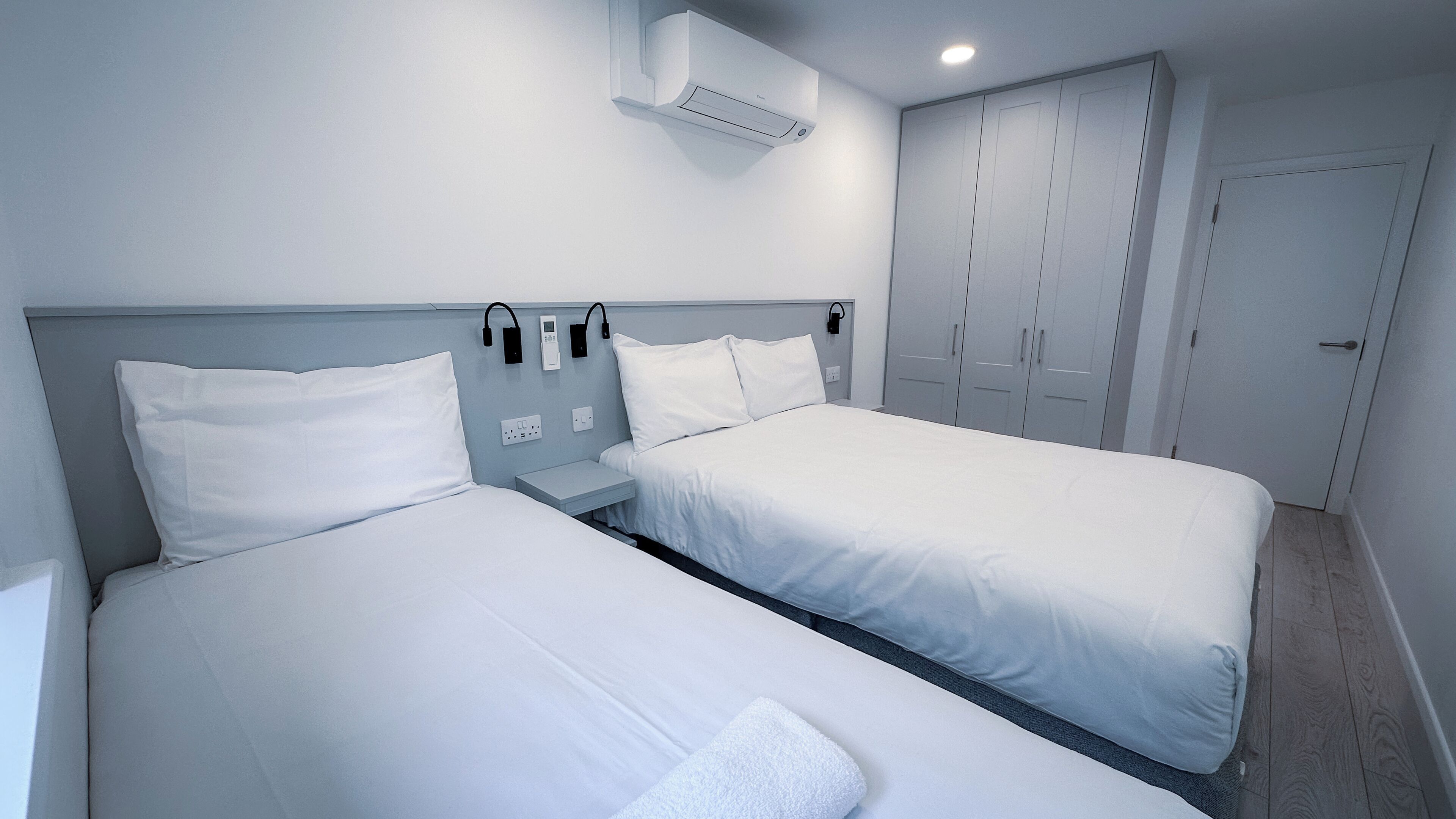A hotel room with two beds, a wardrobe, and a ceiling-mounted air conditioner.