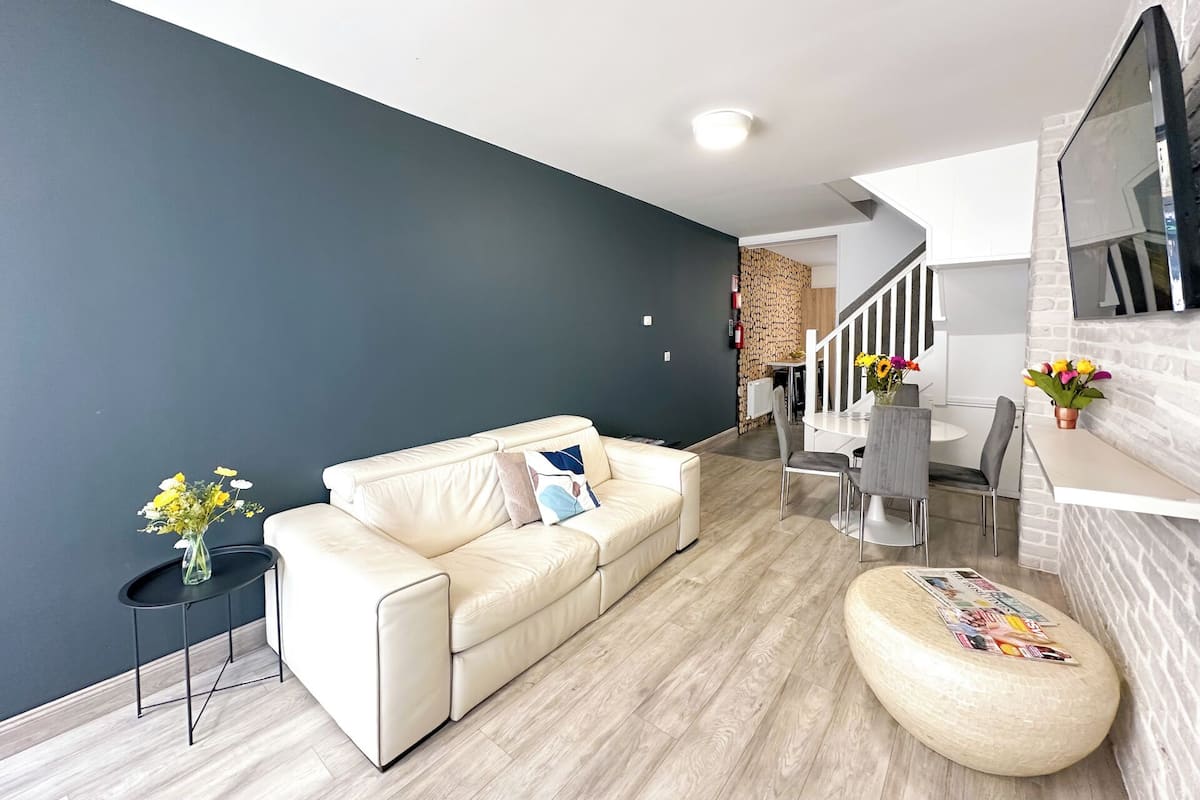 Een moderne woonkamer met een donkere wand, een beige bank, een rond hocker en een eethoek met een witte tafel en stoelen.