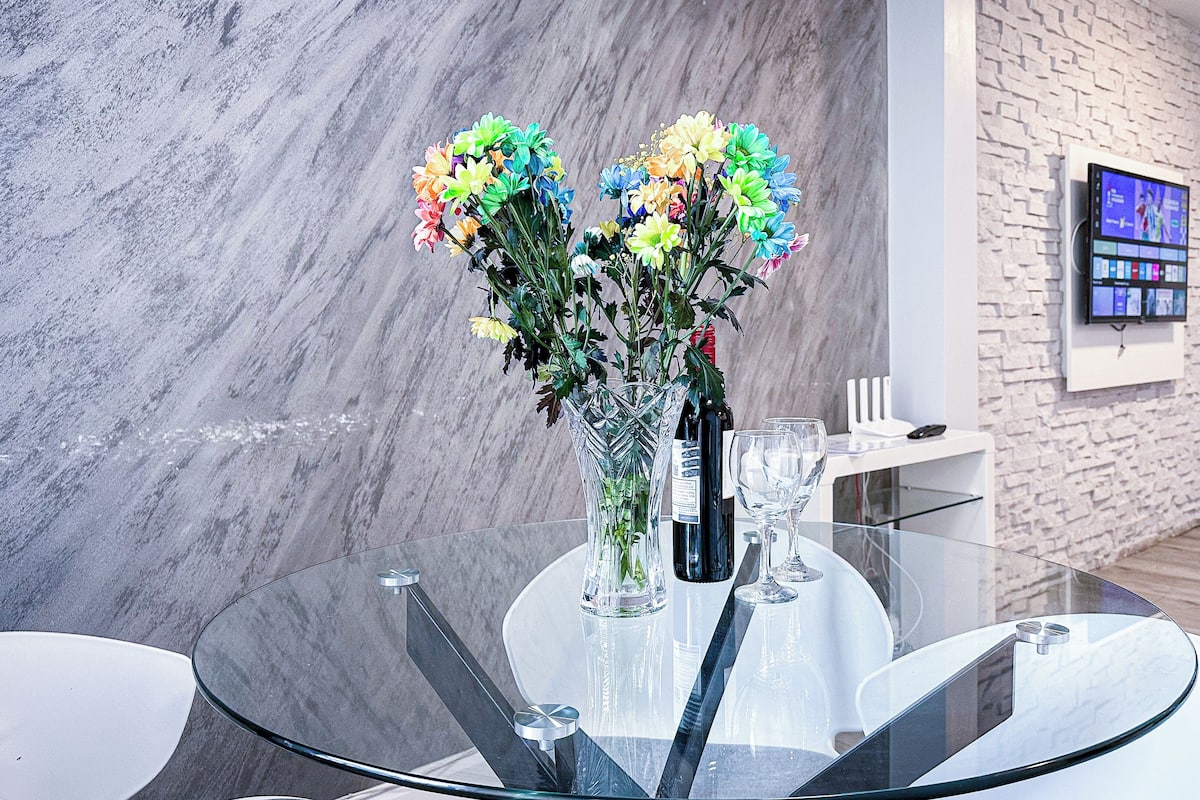 Een modern interieur met een glazen tafel, een vaas met kleurrijke bloemen, een fles wijn en een televisie aan de muur.