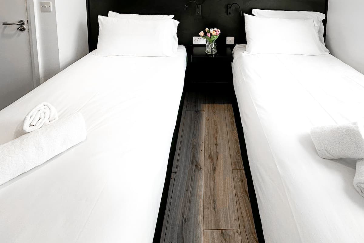 Een hotelkamer met twee eenpersoonsbedden, elk met een witte deken en een opgerolde witte handdoek. Een nachtkastje met een bloevas staat tussen de bedden.