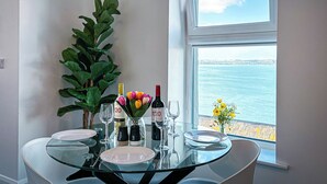 Una mesa de comedor con cubierta de vidrio, rodeada de sillas, dispuesta con platos, copas de vino y un florero. Un ventanal ofrece vistas al mar.