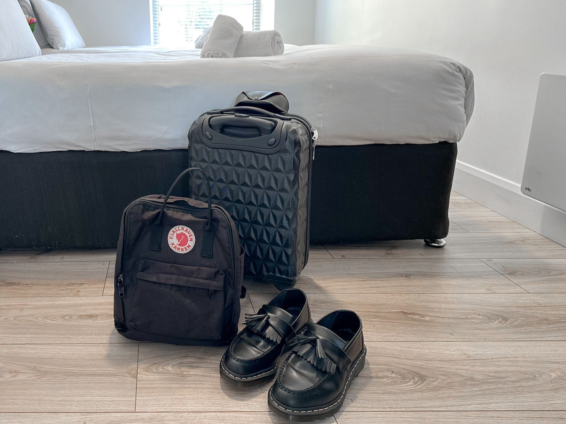 A bed with a grey headboard, a grey suitcase, a black bag with a logo, and a pair of black shoes on a wooden floor.