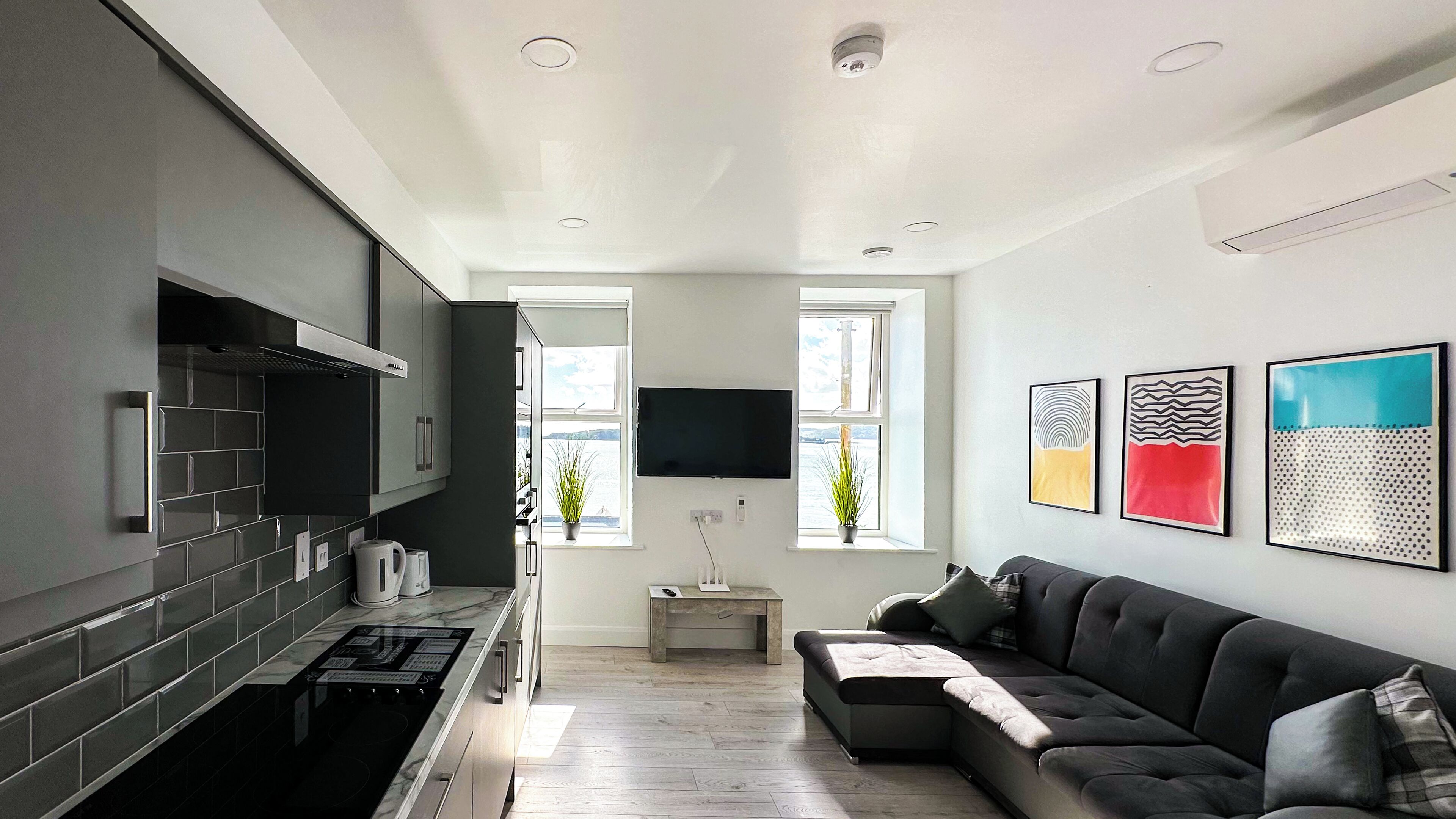A modern kitchen with a built-in stove, a dining area with a table and chairs, and a living room with a sofa and a flat-screen TV mounted on the wall.
