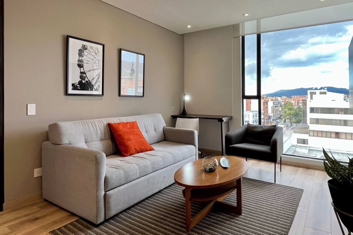 Una sala de estar moderna con un sofá gris, una mesa de centro y vistas de un paisaje urbano a través de amplias ventanas.