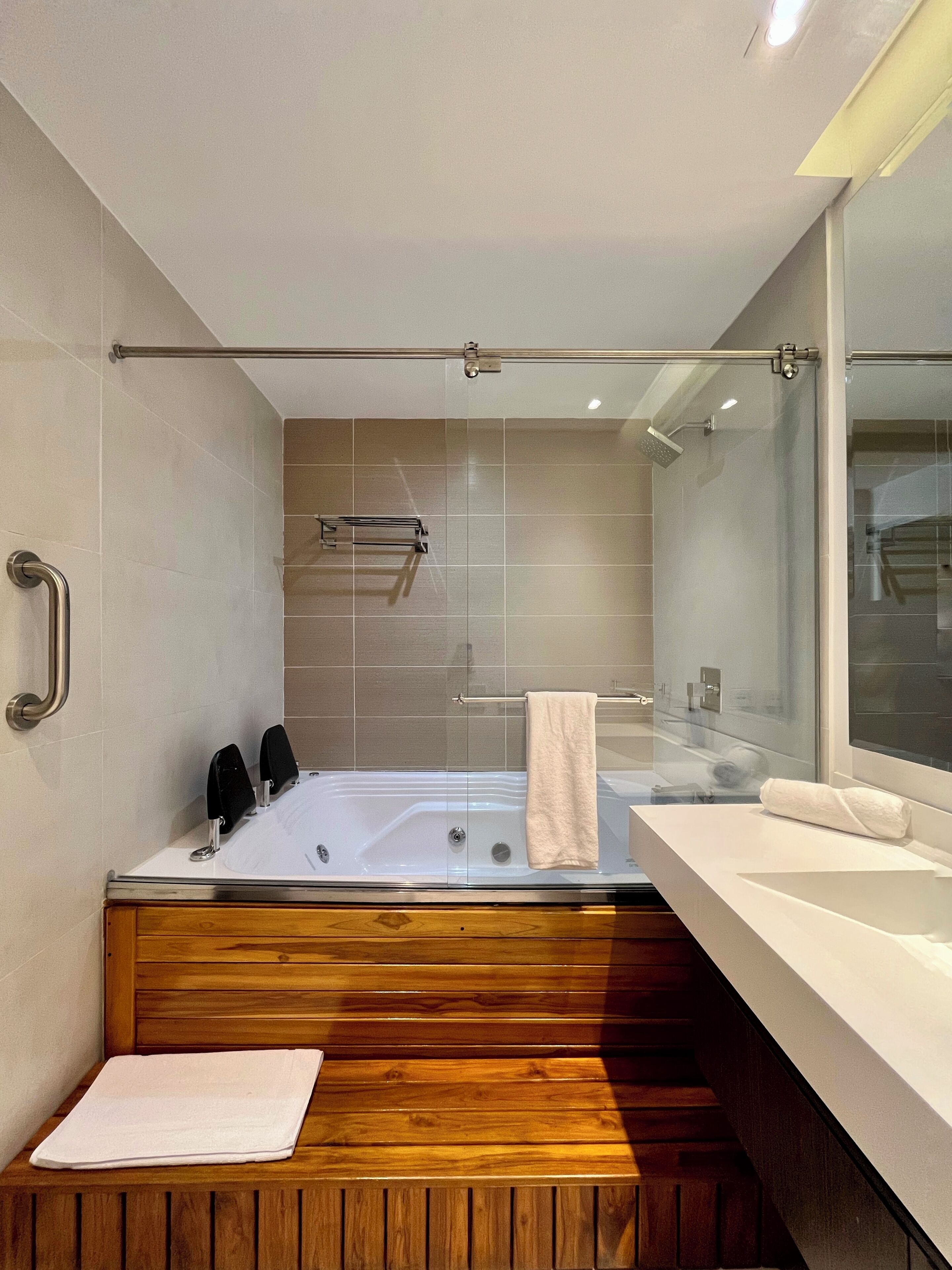 Un baño moderno con bañera de hidromasaje con mampara de vidrio, banca de madera y un espejo grande.