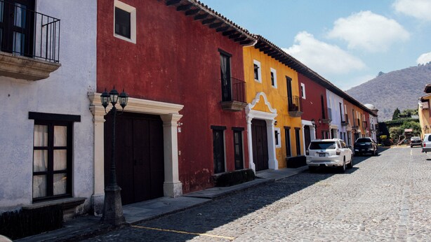 Una calle adoquinada flanqueada por edificios coloridos, un coche blanco estacionado al lado, y una montaña al fondo.