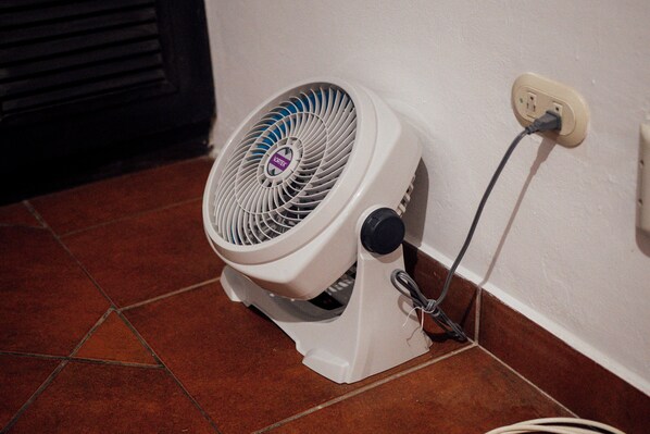 Ventilador portátil apoyado contra una pared, conectado a un enchufe mediante un cable.
