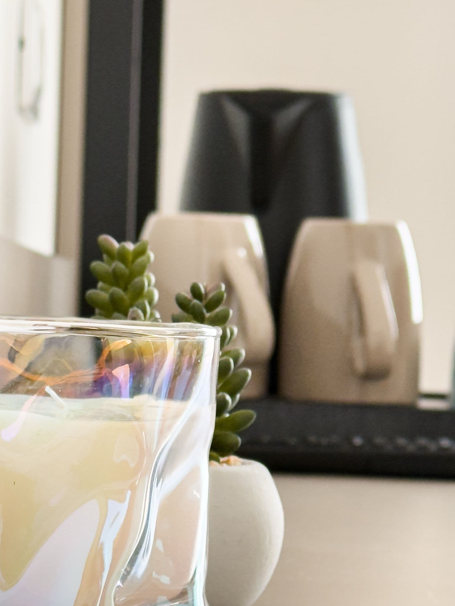 A clear glass candle in a room with a neutral color scheme, featuring a small succulent plant and a couple of beige mugs in the background.