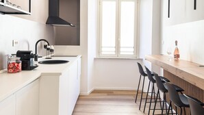 Una cocina moderna con armarios blancos, una campana extractora negra, un ventanal con persianas y una barra con taburetes.