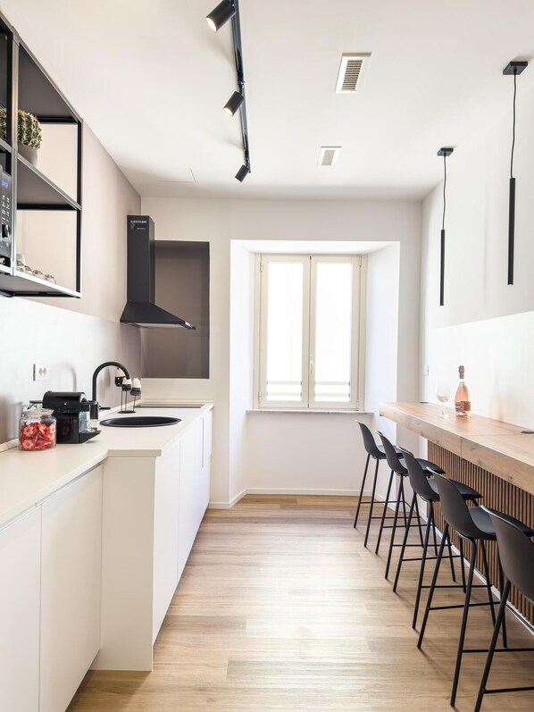 A modern kitchen with white cabinetry, a black range hood, a window with blinds, and a bar area with stools.